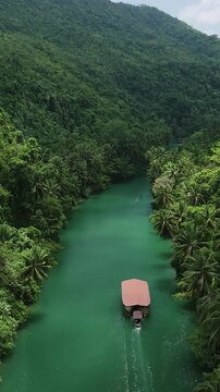 Vertical drone ascends while following a Loboc River cruise boat downstream through green water between forested hills in Bohol
