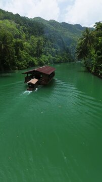 Vertical drone orbits left around a Loboc River cruise boat traveling downstream through green water and forested hills in Bohol