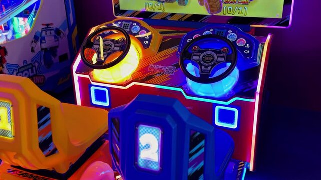 A brightly illuminated dual car racing arcade machine stands empty inside a neon-lit indoor amusement center. The colorful game features glowing yellow and blue steering wheels ready for drivers.