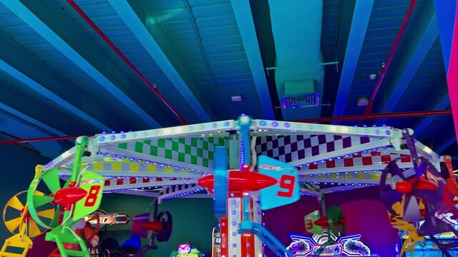 A colorful spinning airplane ride operates inside a bright indoor amusement center. The mechanical attraction features flashing LED lights and checkered patterns under a blue industrial ceiling.