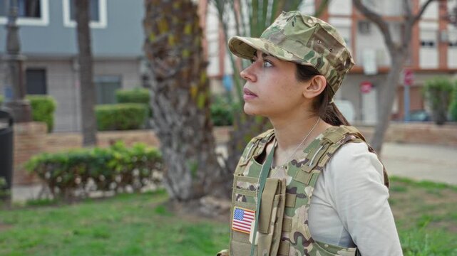 Woman soldier in army uniform and camouflage cap with us flag patch looks up in profile, face visible on street near palm trees and building; pride duty.