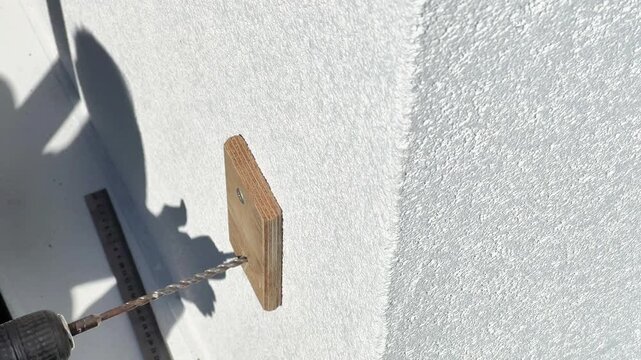 Close-up shot of an electric drill bit drilling through a wooden template into a white plaster wall. Ideal for power tool commercials, DIY guides, and renovation videos.