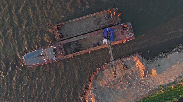 Top down aerial view of industrial barges and a conveyor belt moving sand and soil to reinforce a river dike during a golden sunset