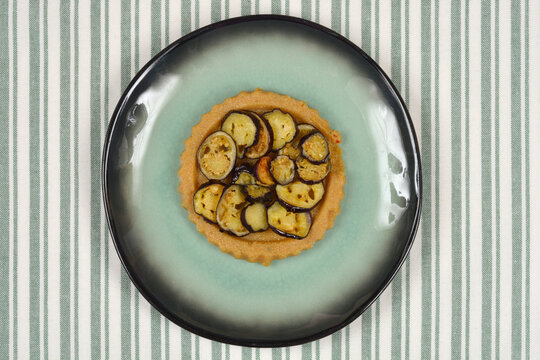 Top view of a savory cornmeal polenta tart topped with fried eggplant slices on a green plate over a striped tablecloth.