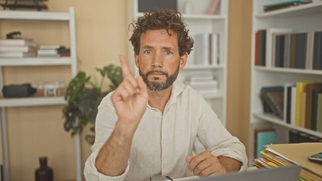 Man points finger to camera on studio desk with laptop, folders, books and plant, making a clear pointing gesture; focus.