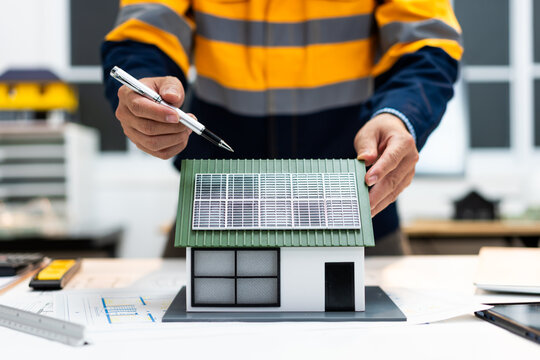 Engineer in high visibility workwear points at a small house model with rooftop solar panels, showing renewable energy planning in a modern office.