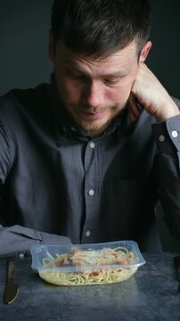 Sad and lonely man sitting at a dark table, staring at a prepackaged pasta meal with a look of disappointment and unhappiness, highlighting feelings of isolation and dissatisfaction. Vertical video