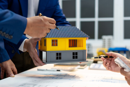 Real estate agents discuss a small house model over building plans, showing home buying, property investment and mortgage planning in a modern office