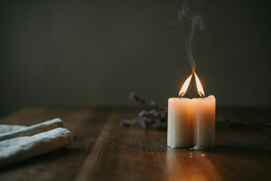 Two white pillar candles burning on a dark wooden table with a soft linen cloth and dried lavender sprigs in a dim, moody interior setting