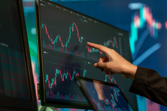 Close-up of a trader pointing at financial candlestick charts on multiple computer screens in a dark office, showing stock market analysis and online investment.