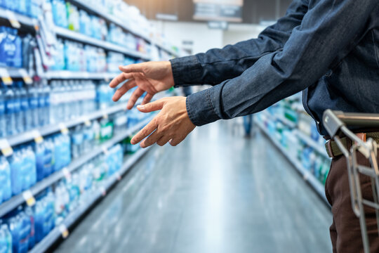Close-up of a shopper reaching toward shelves of bottled water in a supermarket aisle, with a shopping cart and blurred store background.