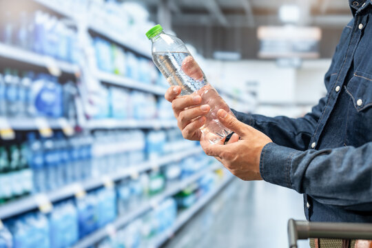 Close-up of a shopper reading a clear plastic water bottle label in a supermarket aisle, choosing bottled water from shelves.