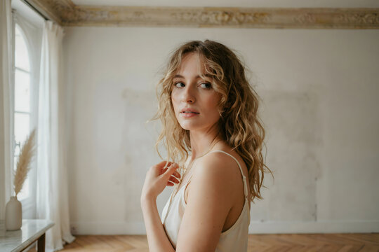 Young woman with wavy blonde hair wearing a beige camisole standing in a bright room with white walls and a window with sheer curtains