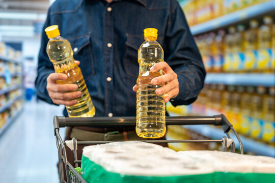 Shopper compares two bottles of cooking oil while standing behind a grocery cart in a supermarket aisle.