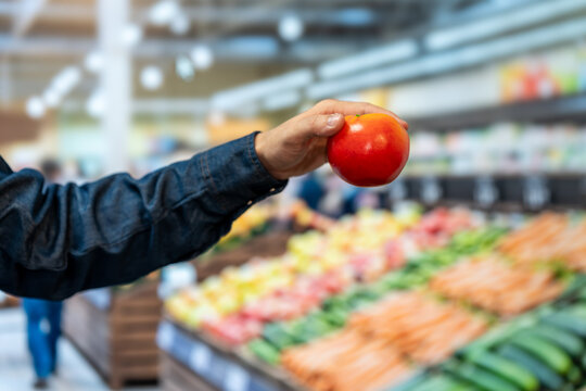 Close-up of a shopper holding a ripe red tomato in a bright supermarket produce aisle, with fresh vegetables softly blurred in the background.