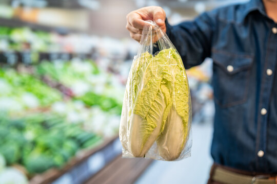 Shopper holding a plastic bag of fresh napa cabbage in a supermarket produce aisle, showing healthy grocery shopping and food packaging.
