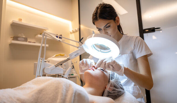Professional cosmetologist using magnifying lamp during manual facial cleaning procedure