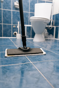 Steam mop sanitizing blue ceramic bathroom tiles near a toilet, using hot vapor to remove dirt, grime and germs for a fresh, sparkling, hygienic floor surface. Concept of household cleanliness.