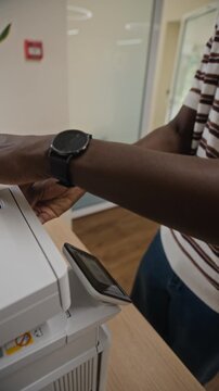 Vertical video of young African American man scanning document on office printer and checking smartwatch. Footage is perfect for corporate training and business productivity commercials