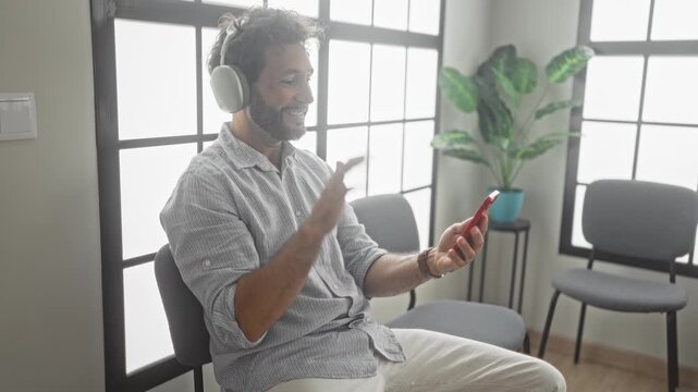 Man holding smartphone and waving hand while seated in chair by frosted windows and potted plant in a building; contentment connection.