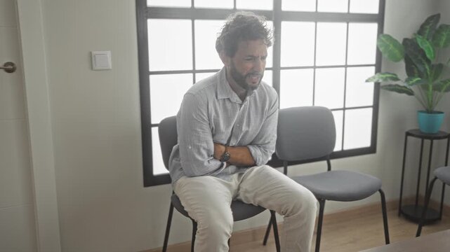 Man sitting on chair with arms crossed and hands clasped over stomach in building waiting area near window and plant; anxiety waiting.