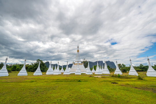 White pagodas of Toh Tae Ploe in Kayin State, Myanmar, set against lush green fields and scenic mountains under a cloudy blue sky with grazing cattle.