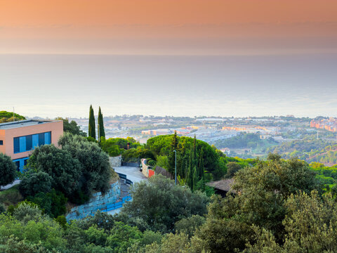 Scenic elevated panoramic view over Mataro coastal landscape at sunset with modern villa nestled in lush green Mediterranean trees and hazy calm sea horizon.