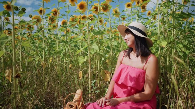 Woman in pink dress sits among sunflowers with picnic items on blanket. Sunflower field under blue sky tall green stems and yellow blooms surround her. Close up view emphasizes relaxed pose