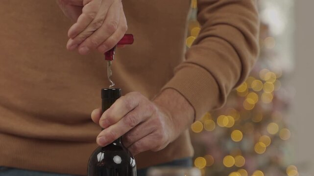 Close-up of a man's hands using a corkscrew to open a bottle of red wine, with festive blurred lights in the background