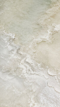 Waves hit the shore, creating a foamy texture on the wet sand. The sun shines bright above, illuminating the scene at midday