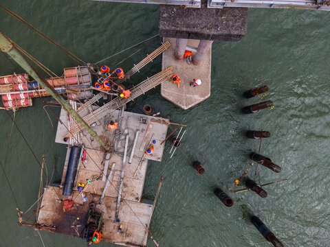 Aerial View of Construction Workers Wearing Orange Safety Jackets Installing Steel Stakes Across a River Beside Large Water Pipeline Infrastructure in Indonesia, Captured from Above.