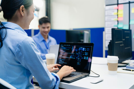 Asian female programmer writing code on a laptop in a modern office with colleague working in background.