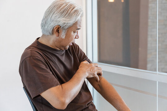 Southeast asian senior man scratching his itchy arm from a mosquito bite or skin rash at home, allergic reaction concept indoor scene
