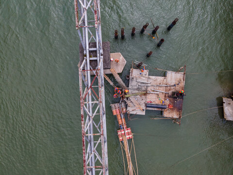 Aerial View of Construction Workers Wearing Orange Safety Jackets Installing Steel Stakes Across a River Beside Large Water Pipeline Infrastructure in Indonesia, Captured from Above.