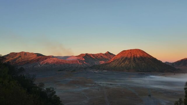 Timelapse from the Tengger Caldera rim showing Mount Bromo scenery transforming from morning twilight to vibrant red sunrise light and bright morning sunshine, Indonesia