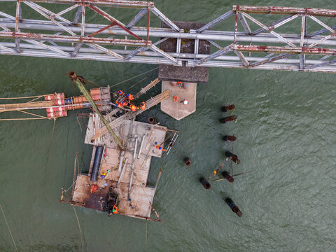 Aerial View of Construction Workers Wearing Orange Safety Jackets Installing Steel Stakes Across a River Beside Large Water Pipeline Infrastructure in Indonesia, Captured from Above.