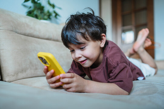 Smiling boy uses a phone while lying on a sofa at home