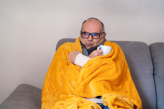 Man wrapped in blanket with hot drink feeling cold in living room