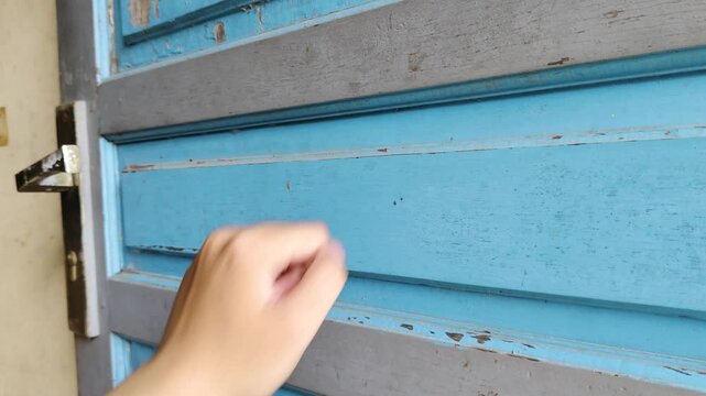 A close-up shot of a hand knocking on a weathered, dark blue wooden door&mdash;with peeling paint and rusty hinges&mdash;in natural light.
