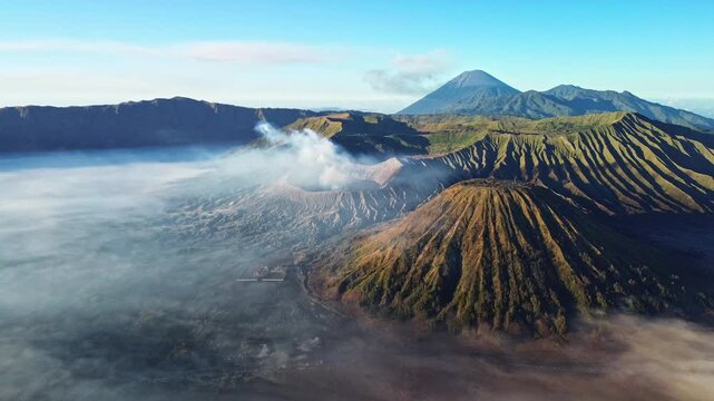 High drone pan across the Mount Bromo volcanic landscape, revealing the Tengger caldera, smoking Bromo crater, Mount Semeru and a sea of clouds beneath a vivid blue sky, Indonesia