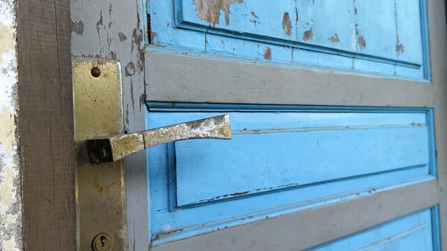 A close-up shot of a hand knocking on a weathered, dark blue wooden door with peeling paint and rusty hinges in natural light.