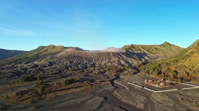 Low drone approach across the Sea of Sand toward Mount Bromo crater rim, passing the Hindu temple and revealing the volcanic landscape beneath a vivid blue sky, Indonesia