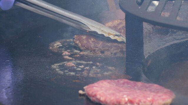 Sizzling burger grilling, Charcoal smoke wafts amid lively evening scene, Crowd gathers eagerly around warm street food event, Juicy beef patties cook on open flame during bustling summer
