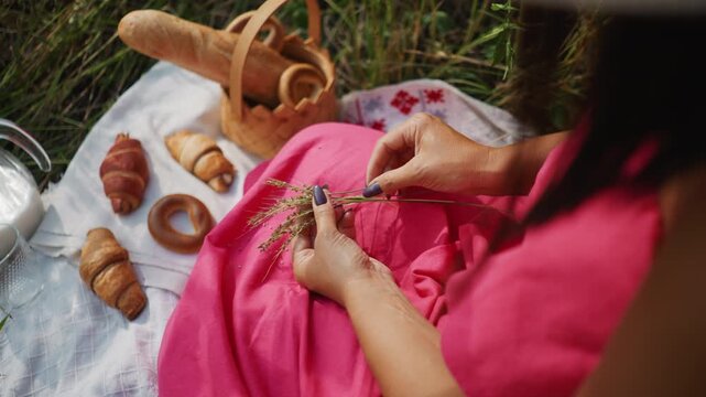Close up of woman in pink fabric arranging wild grass on outdoor picnic blanket with pastries bread and milk. scene features hands manipulating small plants amidst croissants bagels fresh
