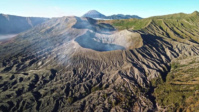 Drone approaches Mount Bromo from the direction of Mount Semeru, revealing the smoking crater interior and rugged volcanic formations beneath a vivid blue sky, Indonesia