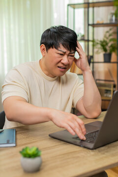 Asian man sitting at home table rereads bad email on laptop, angrily tapping keyboard, brow deeply furrowed. Chinese guy shows tense frustration, shoulders rigid, disappointment and stress visible.
