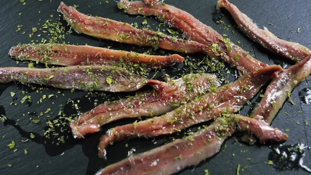 Marinated anchovy fillets sprinkled with chopped herbs arranged on a dark slate surface.