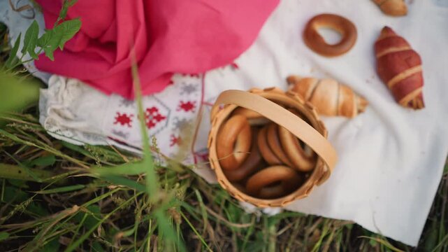 Close up of hand reaching into frame to pick bread from woven basket on white cloth with pink fabric and grass background. Basket contains bagels croissants various baked goods arranged