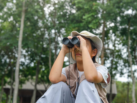 Cute Asian girl in safari hat looking through binoculars outdoors in forest