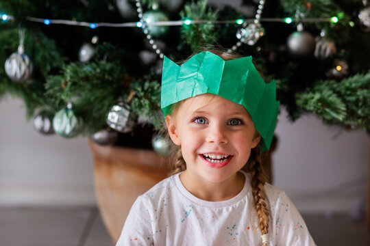 Australian child sitting under Christmas tree wearing green paper cracker hat and smiling